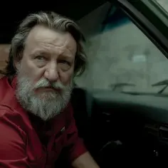 Robert Więckiewicz w czerwonej koszuli, z siwą brodą, siedzi w samochodzie. Jego spojrzenie sugeruje głęboką refleksję, jak w wielu jego filmach.