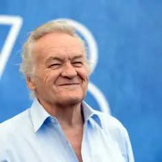 Uśmiechnięty Jerzy Skolimowski, reżyser znany z wyrazistych filmów, na tle niebieskiej kurtyny z białymi symbolami.