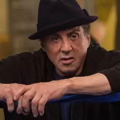 Sylvester Stallone jako Rocky Balboa w czarnym kapeluszu i bluzie, oparty o ring bokserski.