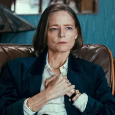 Jodie Foster siedzi w brązowym fotelu, ubrana w białą koszulę i granatową marynarkę.