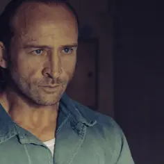 Borys Szyc w filmie, zamyślony, w niebieskiej koszuli. W tle łóżko polowe.
