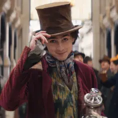 Timothée Chalamet w filmach, jako młody Willy Wonka, w cylindrze i barwnym stroju, z uśmiechem pozdrawia tłum.