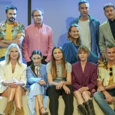 Grupa ludzi pozuje do zdjęcia. Widać obecność obsady, która prezentuje się w różnorodnych strojach, od eleganckich garniturów po luźniejsze, kolorowe ubrania.