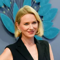Uśmiechnięta Naomi Watts, znana z wielu filmów, w czarnej stylizacji i z kwiatową broszką.