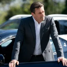 Jon Hamm w garniturze opiera się o czarny samochód, gotowy do kolejnej roli w filmach, serialach i programach.
