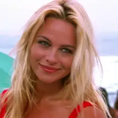 Słoneczny patrol obsada: blond ratowniczka w czerwonym stroju kąpielowym uśmiecha się na plaży, w tle ocean i parasol.