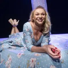 Sonia Bohosiewicz w kwiecistej sukience leży na łóżku. Aktorka znana z wielu filmów uśmiecha się promiennie.