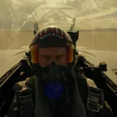 Pilot myśliwca w hełmie z napisem "Maverick" w kokpicie, lecący nad pustynnym krajobrazem.