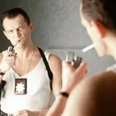 Cezary Pazura w filmach, zapala papierosa przed lustrem. W białej podkoszulce, z odznaką i pistoletem.