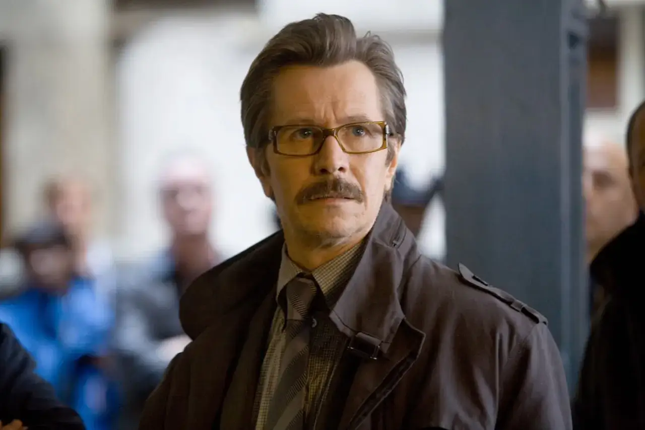 Gary Oldman w okularach i z wąsami, w płaszczu i krawacie. Jego role w filmach i serialach zapadają w pamięć.