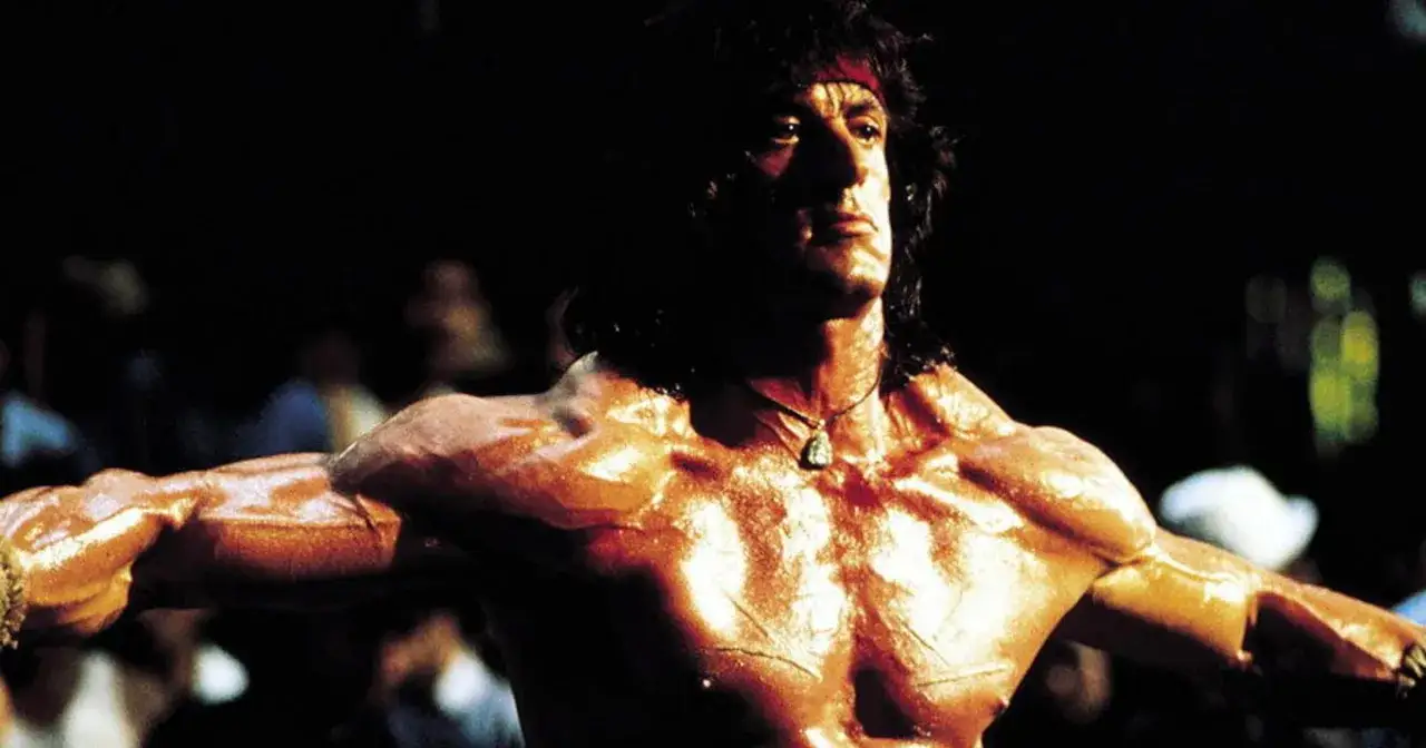 Sylvester Stallone w filmach, z przepoconą klatką piersiową i ramionami, w świetle reflektorów.