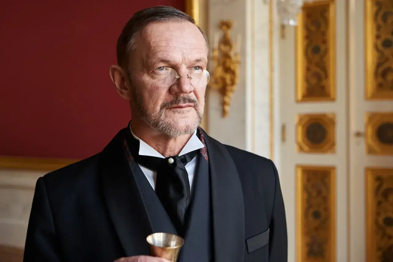 Elegancki mężczyzna w garniturze i okularach, przypominający postać z filmów Cezarego Pazury, trzyma kieliszek.