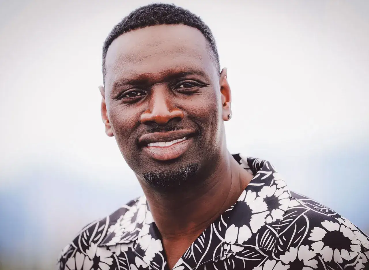 Uśmiechnięty Omar Sy w koszuli w kwiaty. Gwiazda znana z wielu filmów, seriali i programów.