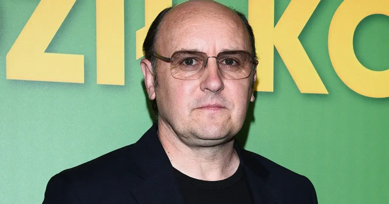 Adam Woronowicz w okularach na tle zielonej ściany z żółtymi literami, przypominający scenę z jego filmów.