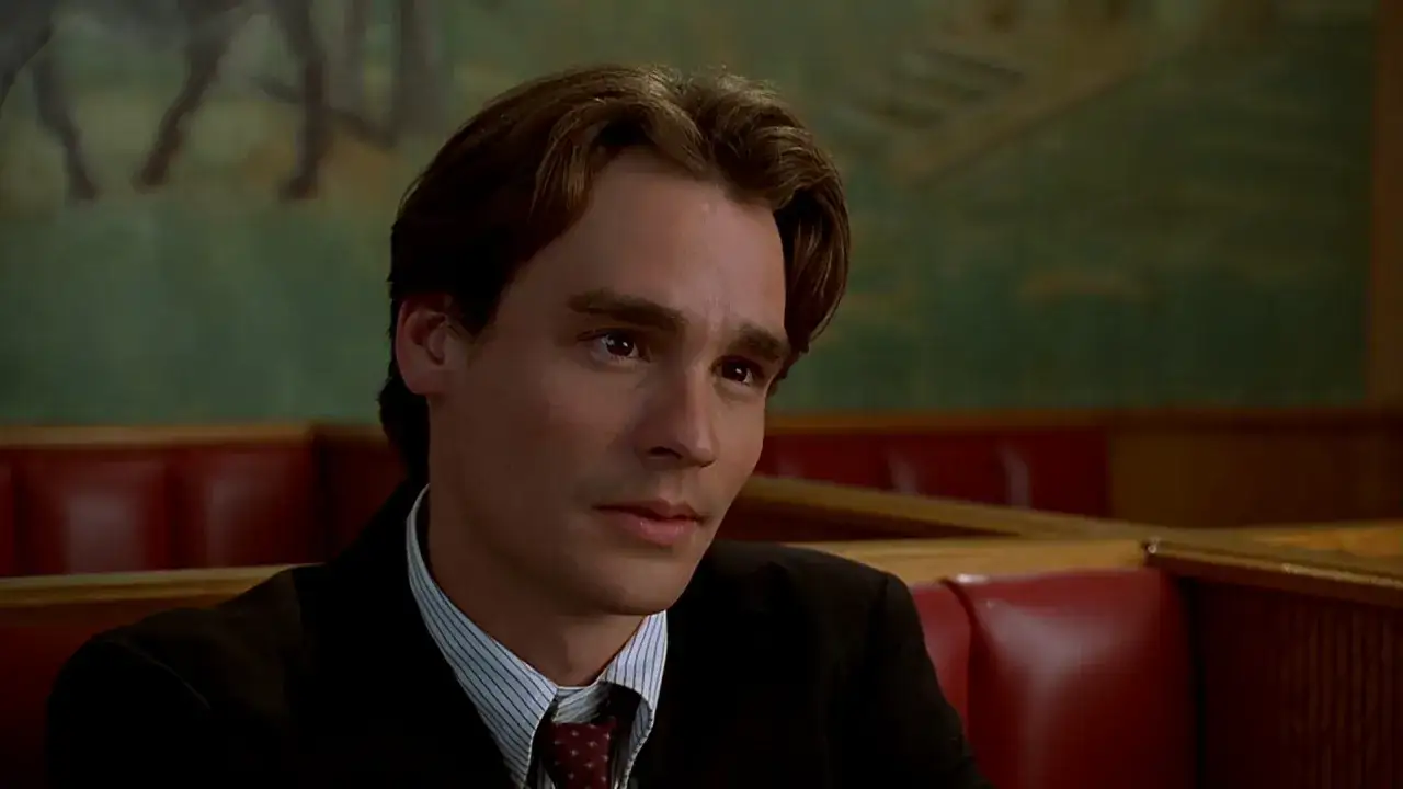 Robert Sean Leonard w garniturze, siedzi w czerwonej kabinie. W tle zielona ściana z malowidłem.