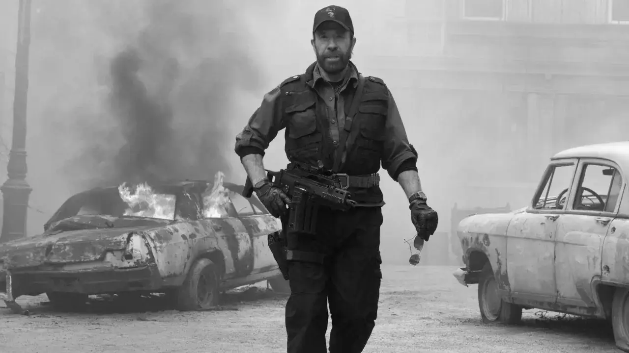 Chuck Norris, mimo swojego wieku, wciąż wygląda na gotowego do akcji. W tle płonący samochód i dym.