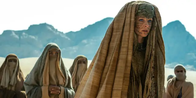 Kobiety w tradycyjnych strojach, w tym jedna z charakterystycznymi tatuażami na twarzy, należące do zakonu **Bene Gesserit**, przemierzają pustynię.